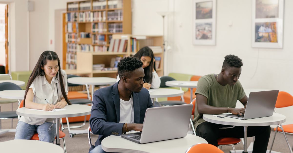 Students working on laptops and notes at a desk in a bright classroom